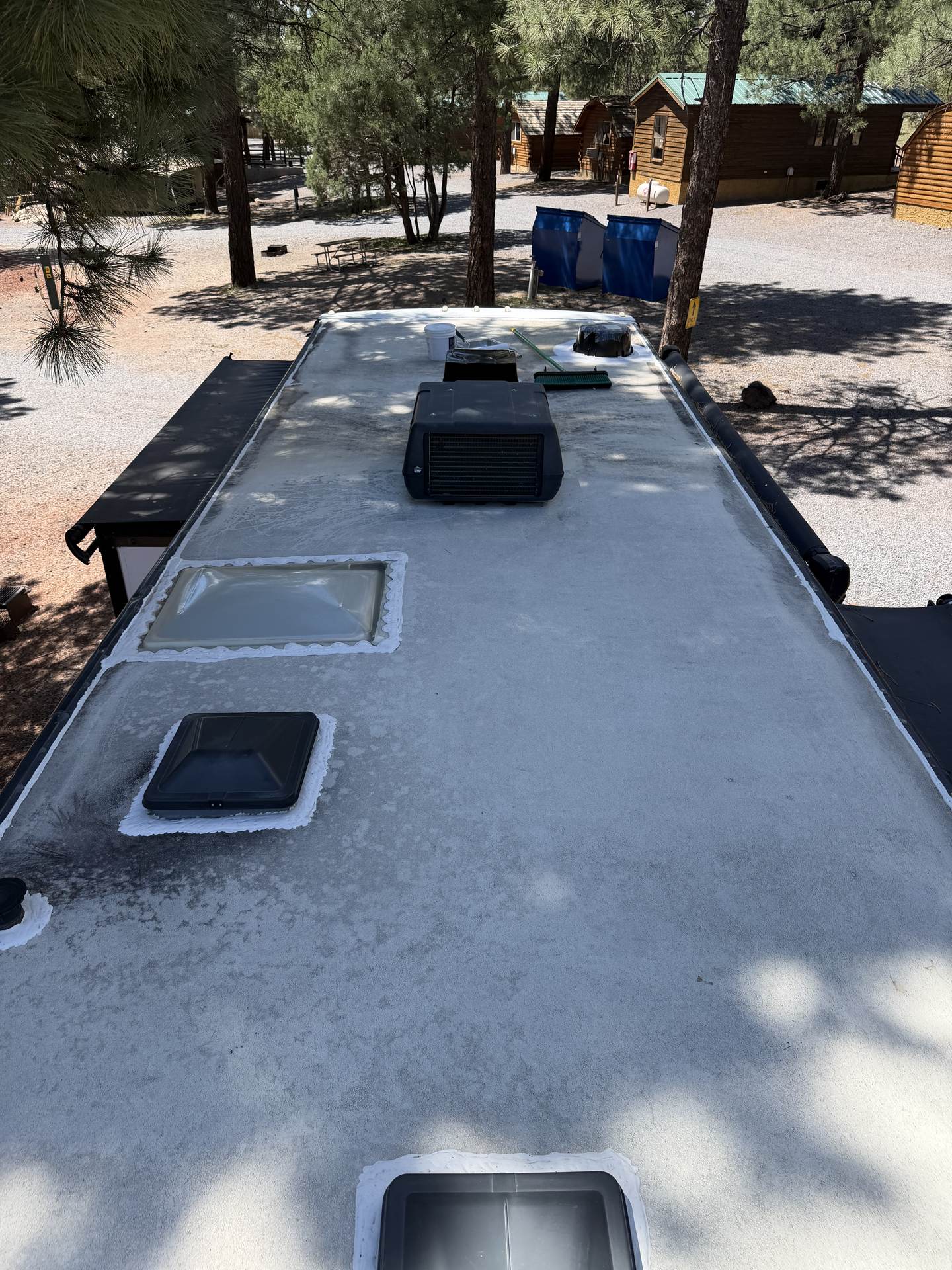 Travel trailer roof mid-reseal with fresh white sealant around the skylight and vents, bucket and brush staged, cabins and pines behind