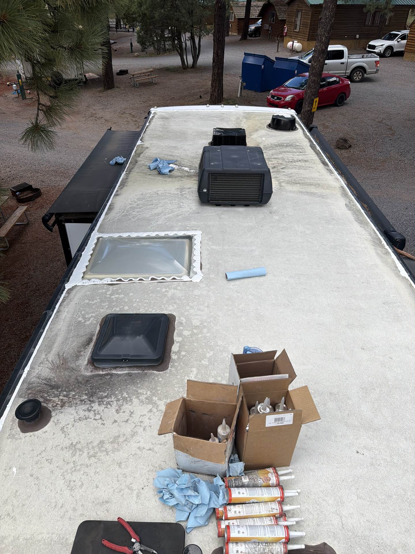RV roof mid-project with the air conditioner shroud set aside, sealant tubes and tools staged, cabins and pines below