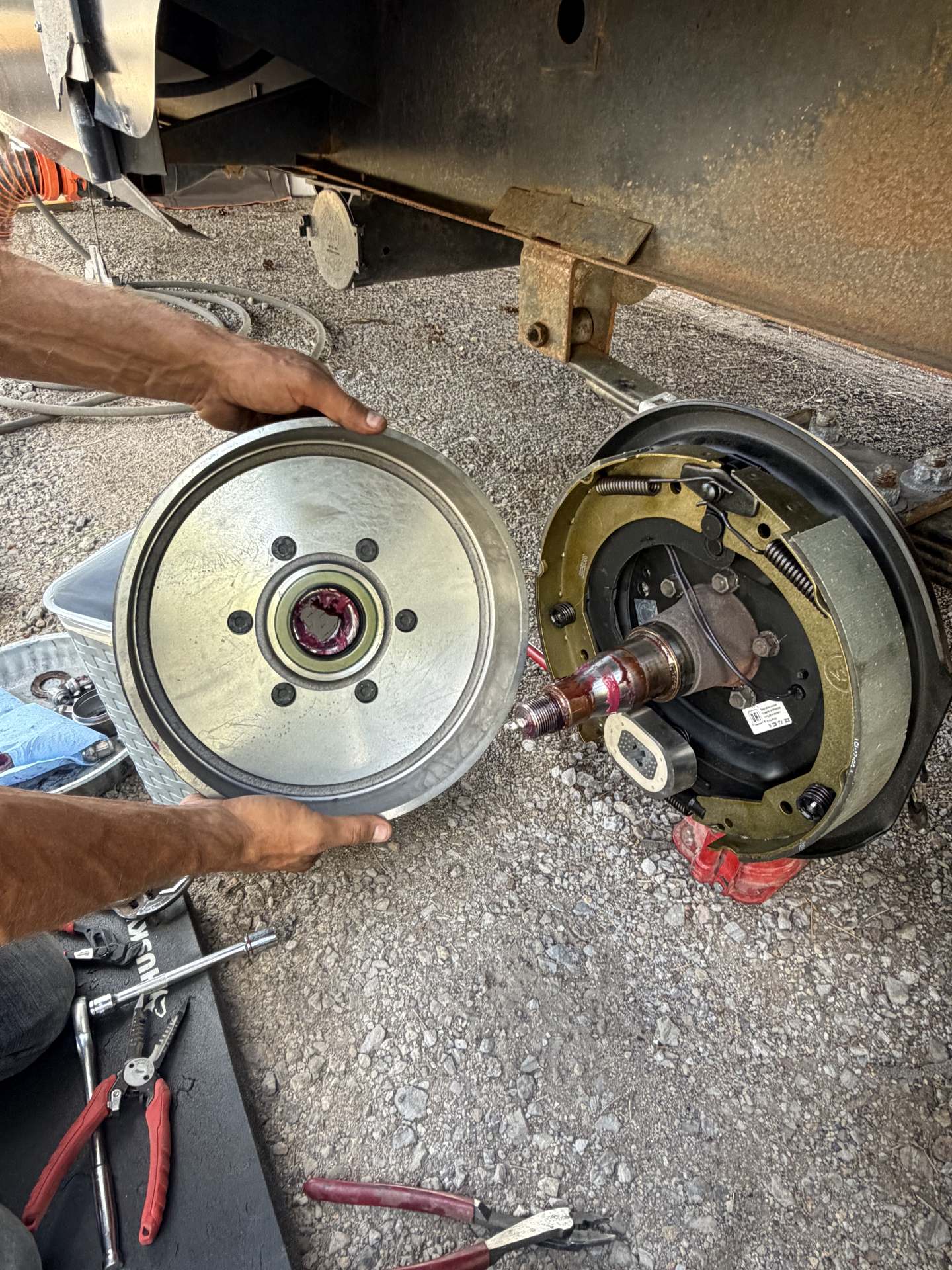 Technician holding an RV brake drum next to a freshly installed trailer brake assembly and greased axle spindle on gravel