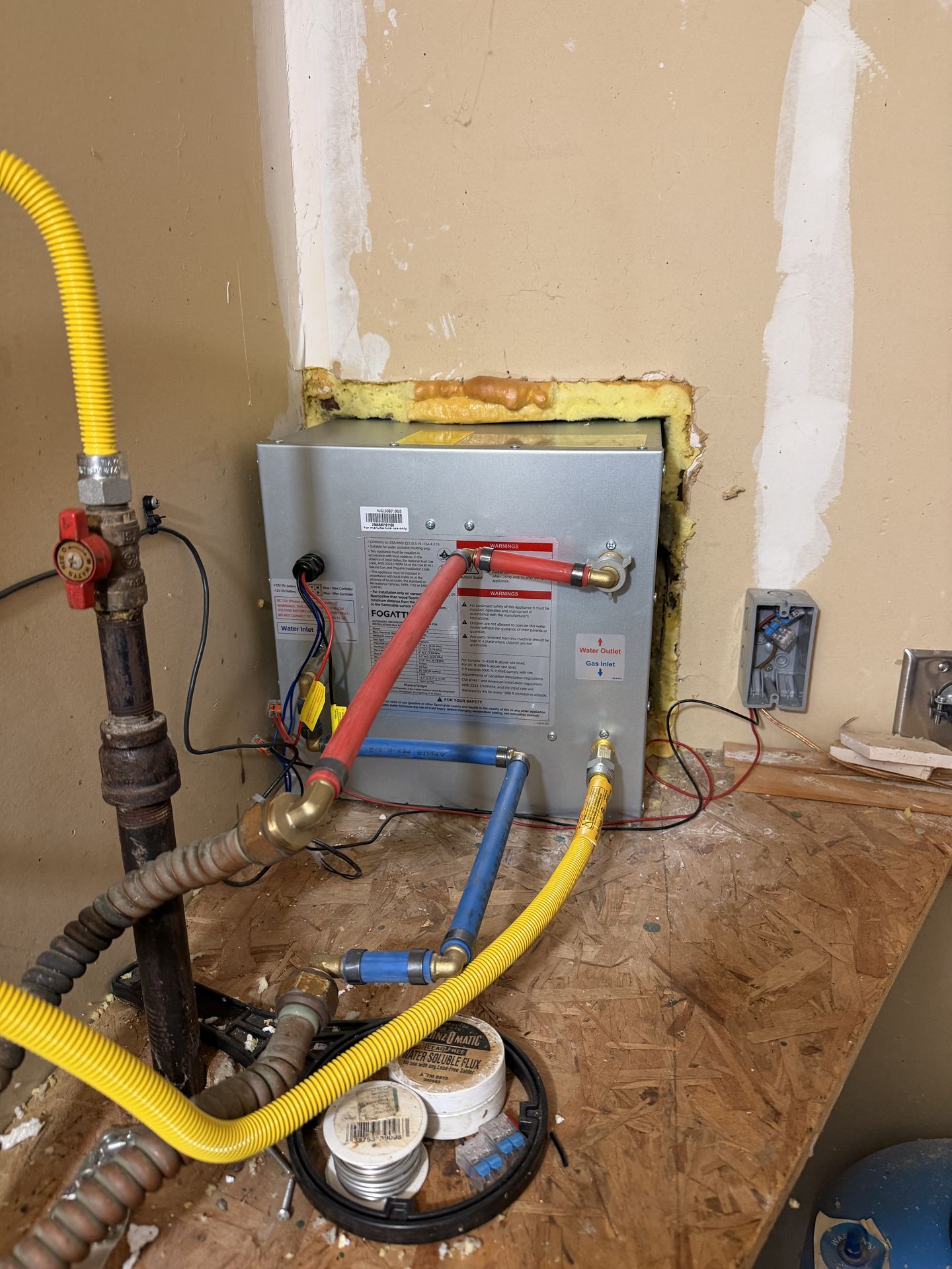 Newly installed RV tankless water heater with PEX water lines, yellow gas flex line, and 12V wiring connected inside the compartment